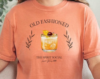 Puede incluir: Una camiseta de color melocotón con un gráfico de un cóctel old fashioned con el texto "Old Fashioned" y "The Spirit Social" impreso en ella.