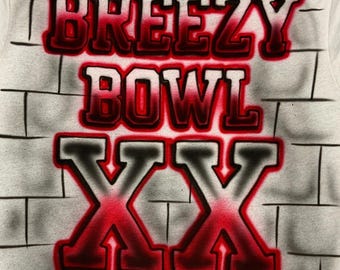 May include: White t-shirt with a brick wall design. The words "BREEZY BOWL XX" are airbrushed in red and black, with a shadow effect. The shirt has a crew neck and short sleeves.