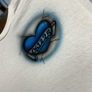 T-shirt premium personnalisé avec impression aérographe — nom en lettres bleues audacieuses avec effet de peinture et date en forme de cœur craquelé