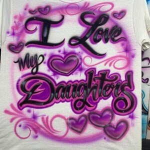 May include: A white short-sleeve t-shirt featuring an airbrushed design in shades of purple and black. The graphic includes the text "I Love My Daughters" in a stylized script font, surrounded by several purple hearts and decorative swirls.