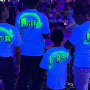 Op de afbeelding: Vier mensen dragen witte t-shirts met neon groene en blauwe ontwerpen. Elk shirt heeft een naam op de rug gedrukt, met een neon groene boog. De shirts worden verlicht door zwart licht, waardoor een gloeiend effect ontstaat.