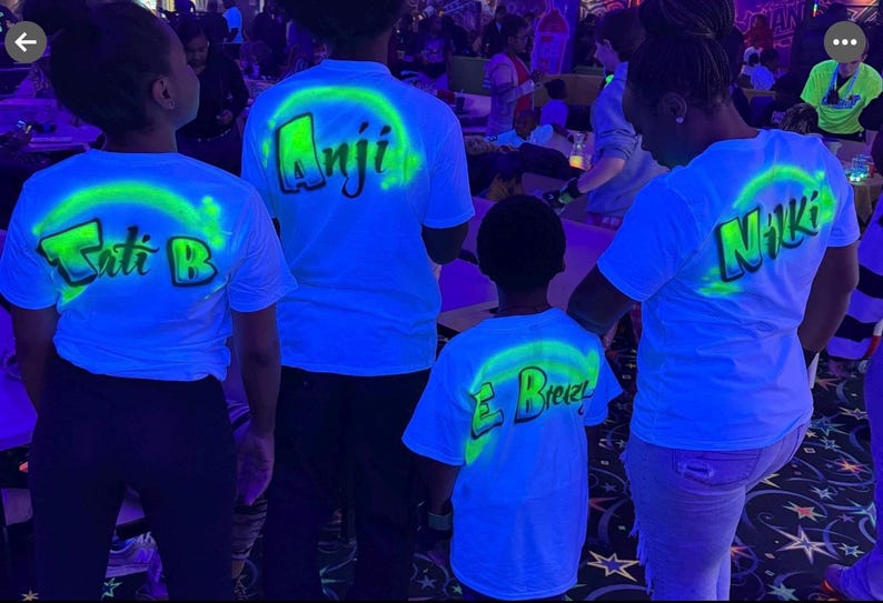 Op de afbeelding: Vier mensen dragen witte t-shirts met neon groene en blauwe ontwerpen. Elk shirt heeft een naam op de rug gedrukt, met een neon groene boog. De shirts worden verlicht door zwart licht, waardoor een gloeiend effect ontstaat.