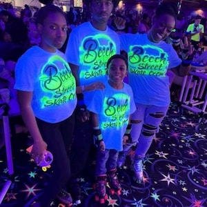 Op de afbeelding: Vier mensen dragen lichtblauwe t-shirts met de tekst "Beech Street Skate Crew" in neon groen. Ze bevinden zich in een rolschaatsbaan, met rolschaatsen en een patroonvloer.