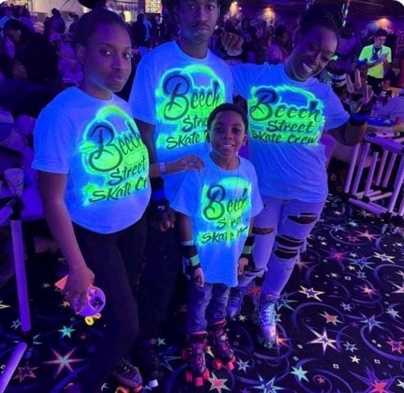 Op de afbeelding: Vier mensen dragen lichtblauwe t-shirts met de tekst "Beech Street Skate Crew" in neon groen. Ze bevinden zich in een rolschaatsbaan, met rolschaatsen en een patroonvloer.