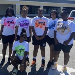Op de afbeelding: Groep mensen die witte t-shirts dragen met gepersonaliseerde namen in verschillende kleuren. De shirts worden gecombineerd met zwarte shorts. De groep staat buiten op een zonnige dag.