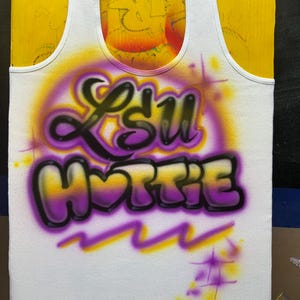 Peut inclure: D&eacute;bardeur blanc avec un motif a&eacute;rographe. Le motif pr&eacute;sente le texte "LSU HOTTY" en violet et noir, avec un fond d&eacute;grad&eacute; jaune et violet. Le d&eacute;bardeur est pr&eacute;sent&eacute; sur une surface jaune.