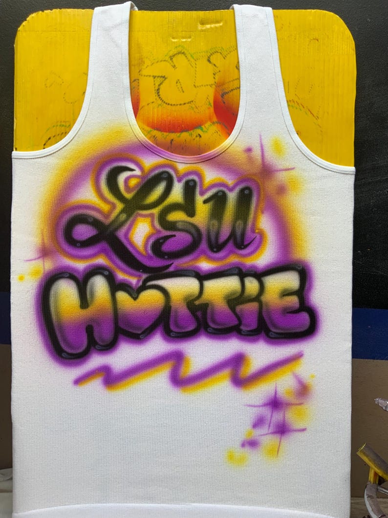 Peut inclure: D&eacute;bardeur blanc avec un motif a&eacute;rographe. Le motif pr&eacute;sente le texte "LSU HOTTY" en violet et noir, avec un fond d&eacute;grad&eacute; jaune et violet. Le d&eacute;bardeur est pr&eacute;sent&eacute; sur une surface jaune.