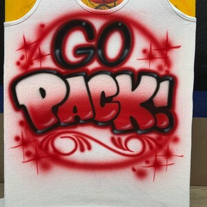 Peut inclure: D&eacute;bardeur blanc avec les mots "GO PACK!" en lettrage a&eacute;rographe rouge et noir. Le motif comprend des &eacute;clats d'&eacute;toiles rouges et des volutes d&eacute;coratives. Le d&eacute;bardeur a une bordure blanche.