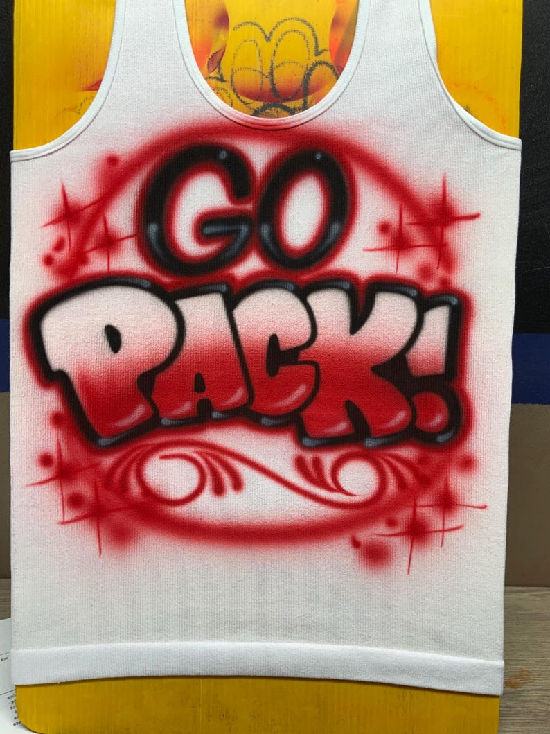 Peut inclure: D&eacute;bardeur blanc avec les mots "GO PACK!" en lettrage a&eacute;rographe rouge et noir. Le motif comprend des &eacute;clats d'&eacute;toiles rouges et des volutes d&eacute;coratives. Le d&eacute;bardeur a une bordure blanche.