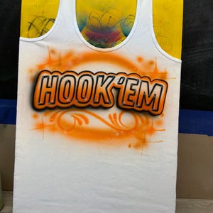 Peut inclure: D&eacute;bardeur blanc avec le texte "HOOK 'EM" en orange et noir, avec un motif a&eacute;rographe orange. Le d&eacute;bardeur est pr&eacute;sent&eacute; sur une forme jaune.