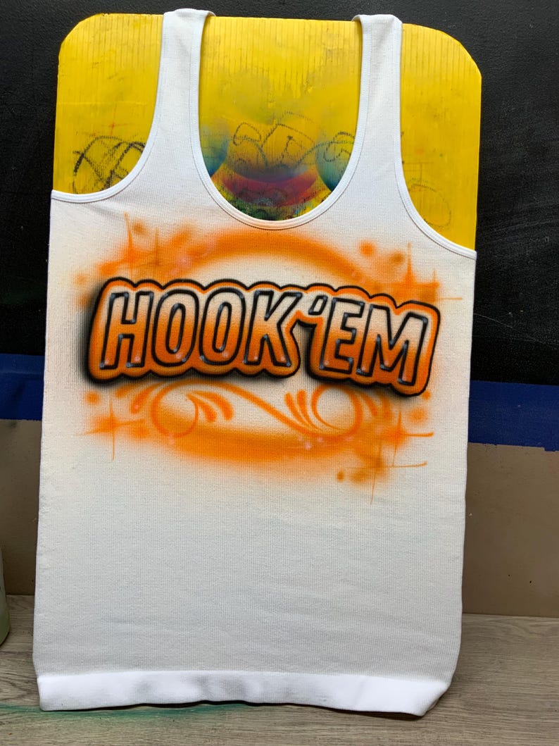 Peut inclure: D&eacute;bardeur blanc avec le texte "HOOK 'EM" en orange et noir, avec un motif a&eacute;rographe orange. Le d&eacute;bardeur est pr&eacute;sent&eacute; sur une forme jaune.