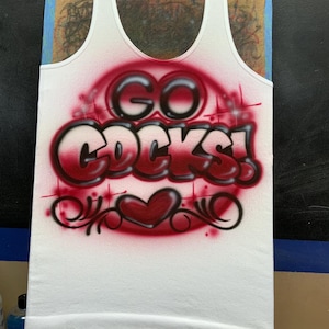 Peut inclure: D&eacute;bardeur blanc avec l'inscription "GO COCKS!" dans un motif rouge a&eacute;rographe. Le motif comprend un c&oelig;ur et des volutes d&eacute;coratives. Le d&eacute;bardeur est pr&eacute;sent&eacute; sur une surface en bois.