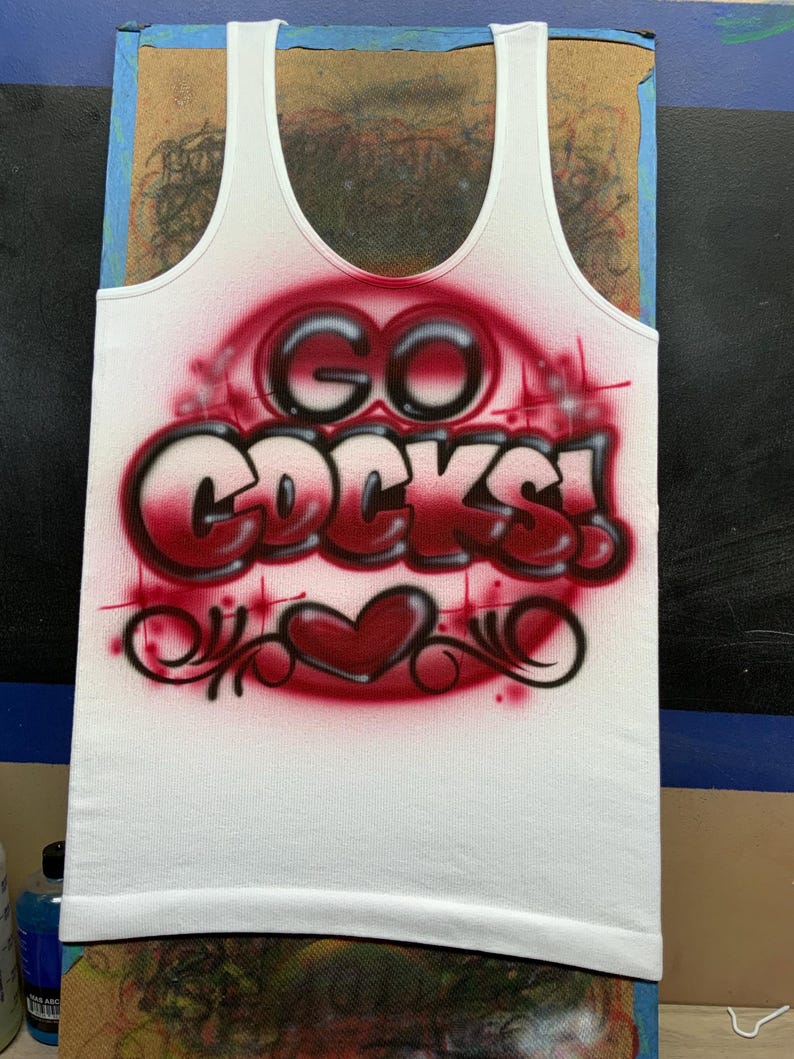 Peut inclure: D&eacute;bardeur blanc avec l'inscription "GO COCKS!" dans un motif rouge a&eacute;rographe. Le motif comprend un c&oelig;ur et des volutes d&eacute;coratives. Le d&eacute;bardeur est pr&eacute;sent&eacute; sur une surface en bois.