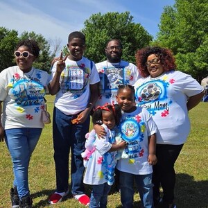 Op de afbeelding: Een groep mensen draagt witte t-shirts met kleurrijke ontwerpen en tekst, staand op een grasveld onder een zonnige hemel. De shirts hebben puzzelstukjesafbeeldingen en de woorden "Autism Mom", "Autism Sister" en "There's a Hero".