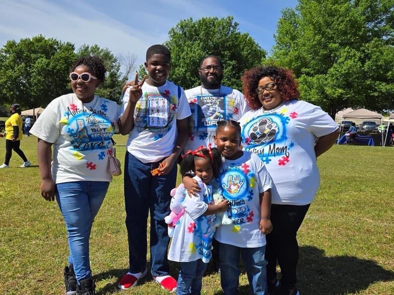 Op de afbeelding: Een groep mensen draagt witte t-shirts met kleurrijke ontwerpen en tekst, staand op een grasveld onder een zonnige hemel. De shirts hebben puzzelstukjesafbeeldingen en de woorden "Autism Mom", "Autism Sister" en "There's a Hero".