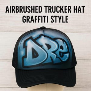 Peut inclure: Casquette de camionneur noire avec un dos en filet et une visière incurvée. Le panneau avant présente des lettres de style graffiti aérographe en nuances de bleu. Le texte "AIRBRUSHED TRUCKER HAT GRAFFITI STYLE" est affiché.