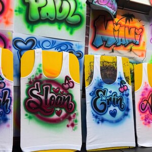 Op de afbeelding: Witte tanktops met op maat gemaakte airbrush namen in verschillende kleuren. Elke top heeft een andere naam, zoals Brennah, Sloan, Erin, Anyssia en Sabrina, met hart- en kersaccenten. Graffiti-achtige letters.