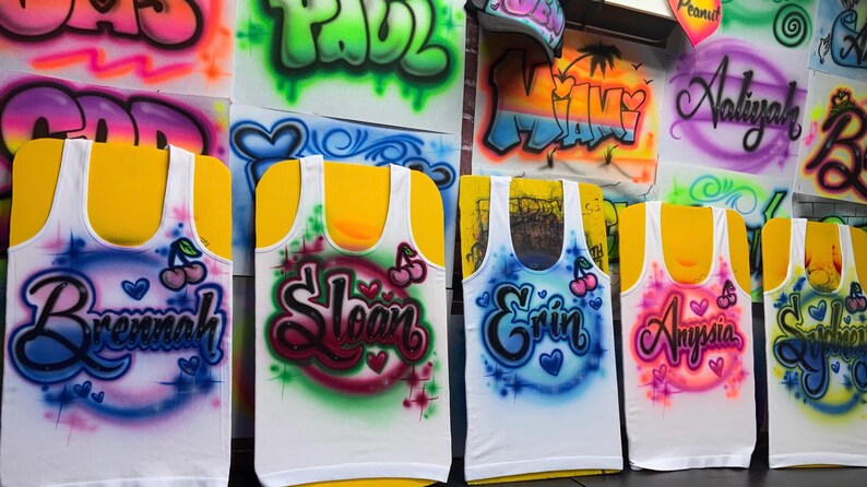 Op de afbeelding: Witte tanktops met op maat gemaakte airbrush namen in verschillende kleuren. Elke top heeft een andere naam, zoals Brennah, Sloan, Erin, Anyssia en Sabrina, met hart- en kersaccenten. Graffiti-achtige letters.