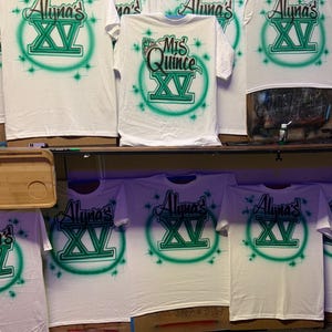 Op de afbeelding: Witte T-shirts met airbrush-ontwerpen. De shirts hebben de tekst "Aluna's XV" in groen en zwart, omringd door een groene cirkel en steraccenten. E&eacute;n shirt heeft "Mis Quince XV". De shirts worden op een plank getoond.
