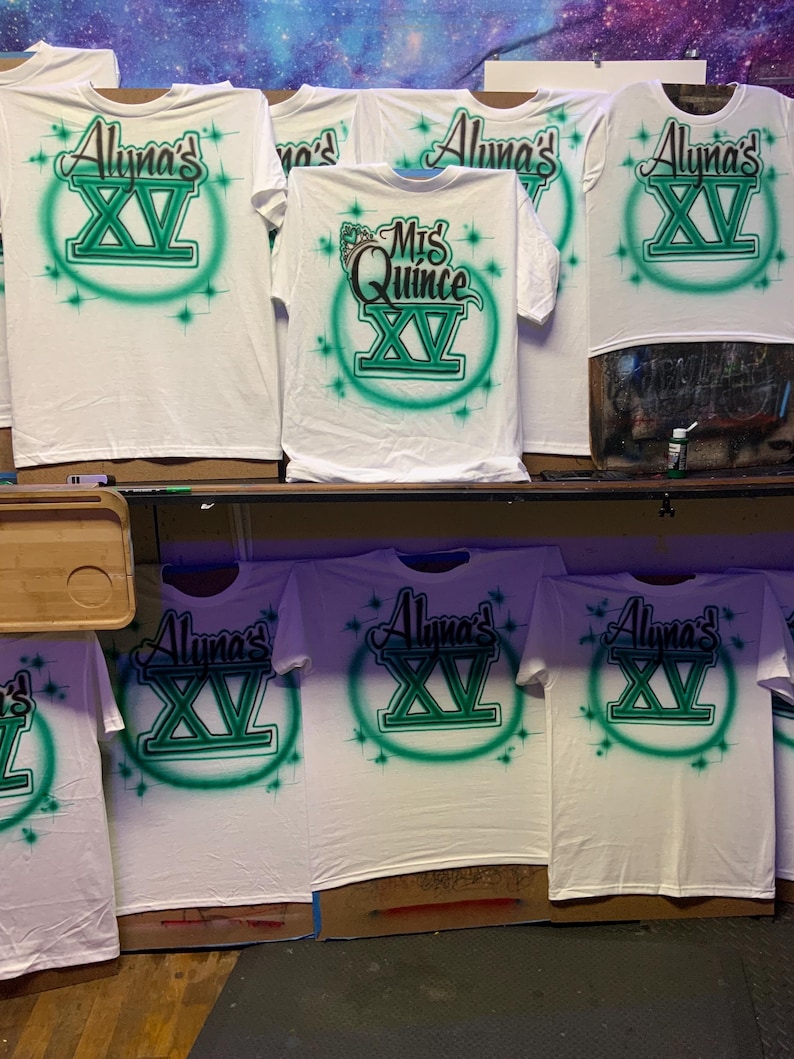 Op de afbeelding: Witte T-shirts met airbrush-ontwerpen. De shirts hebben de tekst "Aluna's XV" in groen en zwart, omringd door een groene cirkel en steraccenten. E&eacute;n shirt heeft "Mis Quince XV". De shirts worden op een plank getoond.