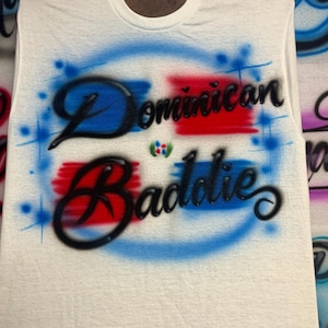 May include: A white sleeveless t-shirt featuring airbrushed blue and red rectangular shapes behind the black cursive text "Dominican Baddie". A small, colorful emblem is positioned between the two lines of text, surrounded by blue starbursts.