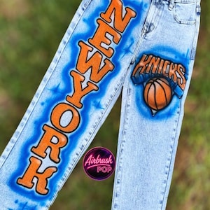 May include: Light-wash denim jeans featuring airbrushed orange and blue designs. The left leg displays the text NEW YORK vertically in orange with blue shading. The right leg features a basketball graphic and the text KNICKS.