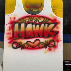 Peut inclure: D&eacute;bardeur blanc avec le nom "HAWK" en lettrage a&eacute;rographe rouge et orange. Le motif comprend un c&oelig;ur et des volutes d&eacute;coratives, le tout sur fond rouge.