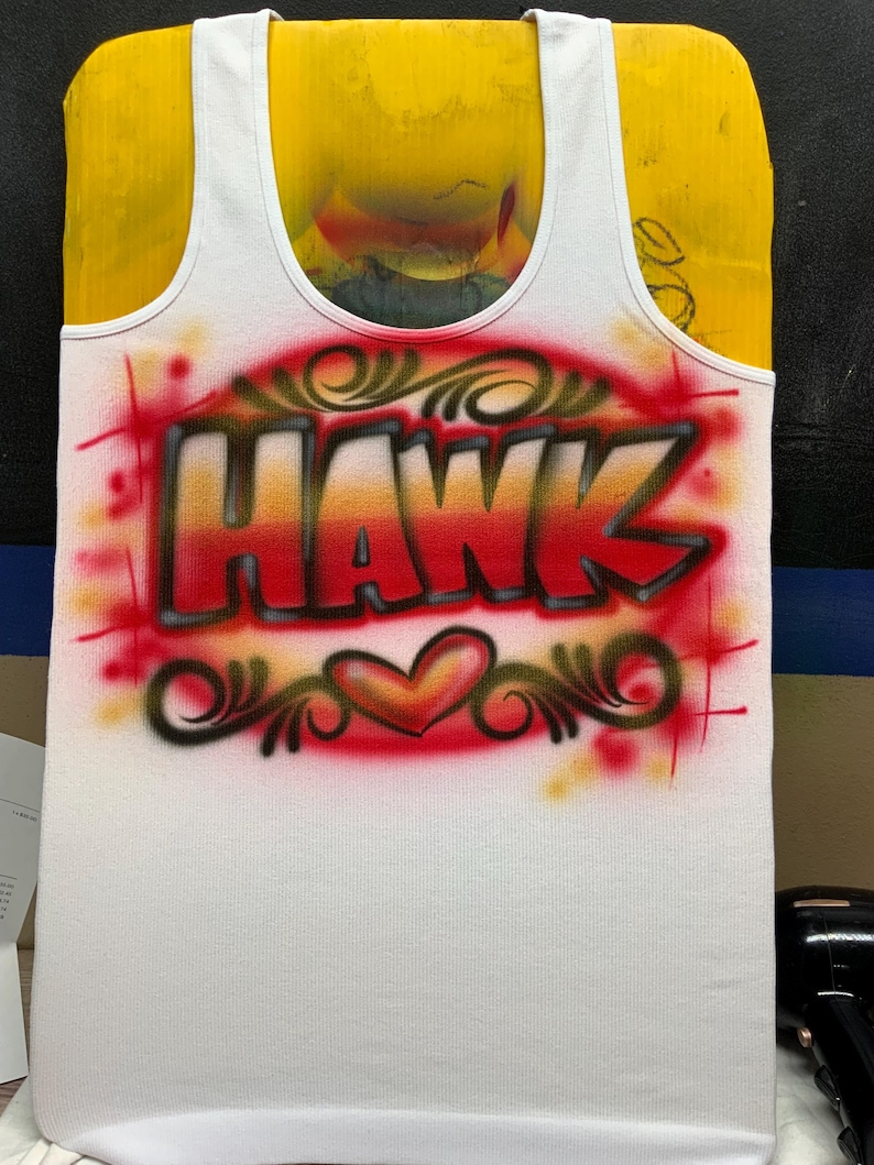 Peut inclure: D&eacute;bardeur blanc avec le nom "HAWK" en lettrage a&eacute;rographe rouge et orange. Le motif comprend un c&oelig;ur et des volutes d&eacute;coratives, le tout sur fond rouge.