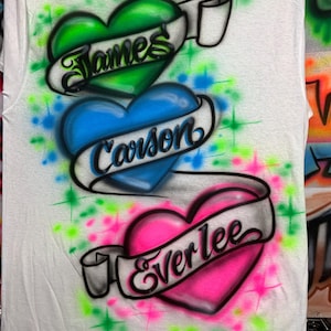 May include: A white t-shirt featuring an airbrushed design of three stacked hearts in green, blue, and pink. Each heart is wrapped in a white banner with black cursive text reading James, Carson, and Everlee, accented by neon starbursts.