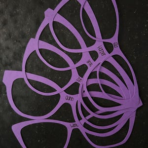 May include: A stack of purple paper eyeglasses cutouts, each with a unique frame design. The names "Harper", "Quinn", "Zuri", "Ella", "Broo", and "Wan" are printed on the frames. The cutouts are arranged on a dark surface.