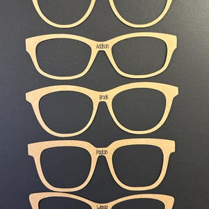 May include: Five pairs of wooden eyeglasses cutouts, each with a different name: Zuri, Addison, Brook, Payton, and Wanda. The frames are a light brown color and are arranged vertically against a dark background. The names are in a simple font.