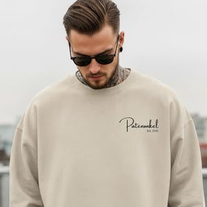 Könnte beinhalten: Beiges Sweatshirt mit dem Wort "Patenonkel" in Schreibschrift, darunter "Est. 2025". Das Sweatshirt hat eine neutrale Farbe und eine lockere Passform. Die Person auf dem Bild trägt eine Sonnenbrille.