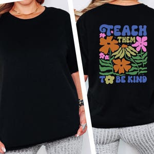 May include: Black t-shirt with the words "TEACH THEM TO BE KIND" in a colorful floral design on the back. The shirt has short sleeves and a crew neck. The front of the shirt is plain black.