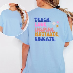 May include: A light blue t-shirt with the words "TEACH. LOVE. INSPIRE. MOTIVATE. EDUCATE." in a retro font on the back. Two small flower graphics are next to the word "INSPIRE". The shirt has short sleeves and a round neck.