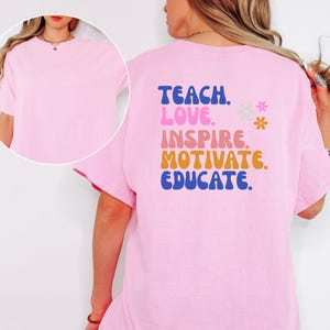 May include: A light pink t-shirt with the words "TEACH. LOVE. INSPIRE. MOTIVATE. EDUCATE." in blue, pink, orange, and yellow lettering. Two small flower graphics are also present. The shirt is a short-sleeved, crew neck style.