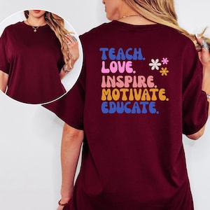 May include: A maroon t-shirt with the words "TEACH. LOVE. INSPIRE. MOTIVATE. EDUCATE." in colorful, retro-style lettering on the back. Two small flower graphics are also present. The shirt has short sleeves and a relaxed fit.