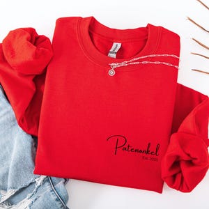 Puede incluir: Una sudadera roja de cuello redondo con el texto negro Patenonkel Est. 2025 impreso en la parte inferior derecha. Un collar de cadena plateada con un peque&ntilde;o colgante circular descansa sobre el cuello de la prenda, combinada con mezclilla azul.