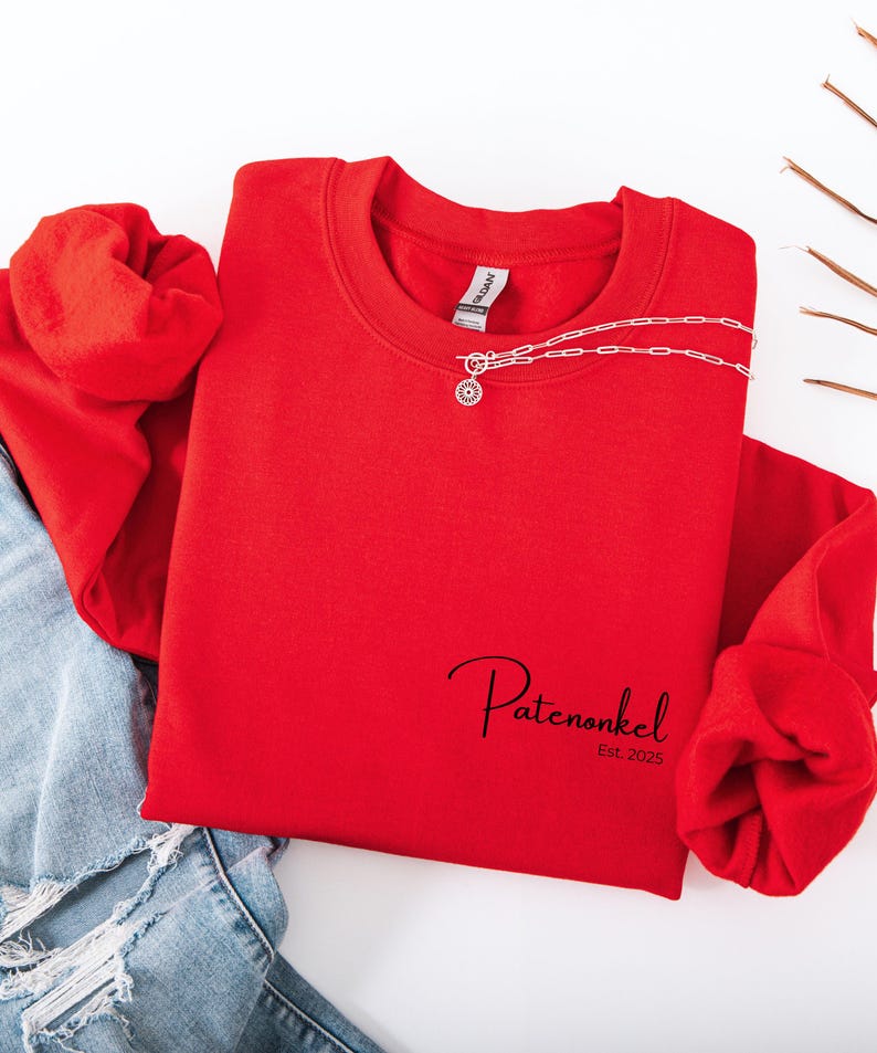Puede incluir: Una sudadera roja de cuello redondo con el texto negro Patenonkel Est. 2025 impreso en la parte inferior derecha. Un collar de cadena plateada con un peque&ntilde;o colgante circular descansa sobre el cuello de la prenda, combinada con mezclilla azul.