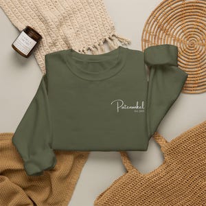Puede incluir: Una sudadera de cuello redondo color verde oliva con texto blanco en cursiva en el pecho izquierdo que dice Patenonkel y un texto m&aacute;s peque&ntilde;o debajo que dice Est. 2025. La prenda tiene mangas largas, cuello y pu&ntilde;os acanalados.