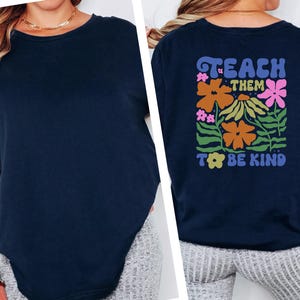 May include: Navy blue crew neck t-shirt. The back of the shirt features the text "TEACH THEM TO BE KIND" with a floral design in orange, pink, and green. The shirt is made of a soft material.