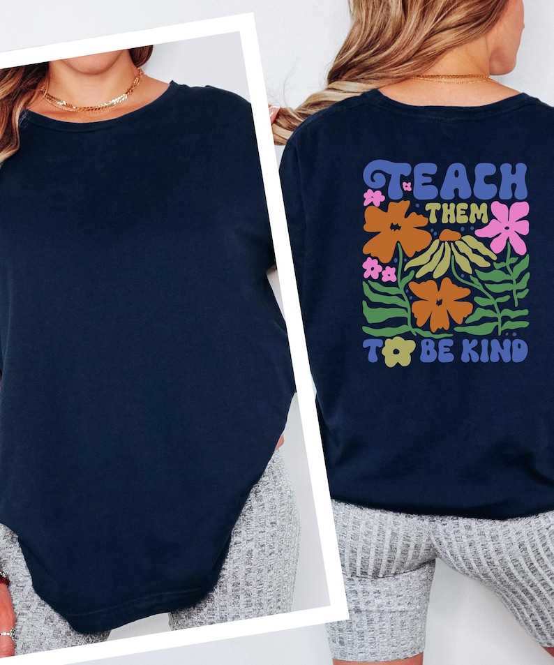 May include: Navy blue crew neck t-shirt. The back of the shirt features the text "TEACH THEM TO BE KIND" with a floral design in orange, pink, and green. The shirt is made of a soft material.