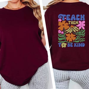 May include: Burgundy-colored long-sleeve shirt with the text "TEACH THEM TO BE KIND" on the back, surrounded by colorful floral graphics. The shirt is a solid color on the front.