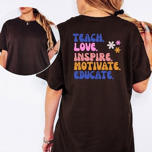 May include: A dark brown t-shirt with the words "TEACH. LOVE. INSPIRE. MOTIVATE. EDUCATE." in colorful text on the back. Two small flower graphics are also present. The shirt has short sleeves and a round neckline.