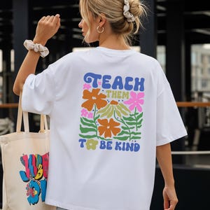 May include: White t-shirt with the text "TEACH THEM TO BE KIND" in blue, orange, and pink floral design. A tote bag with a colorful graphic is held in the left hand. The person is wearing a scrunchie and hoop earrings.