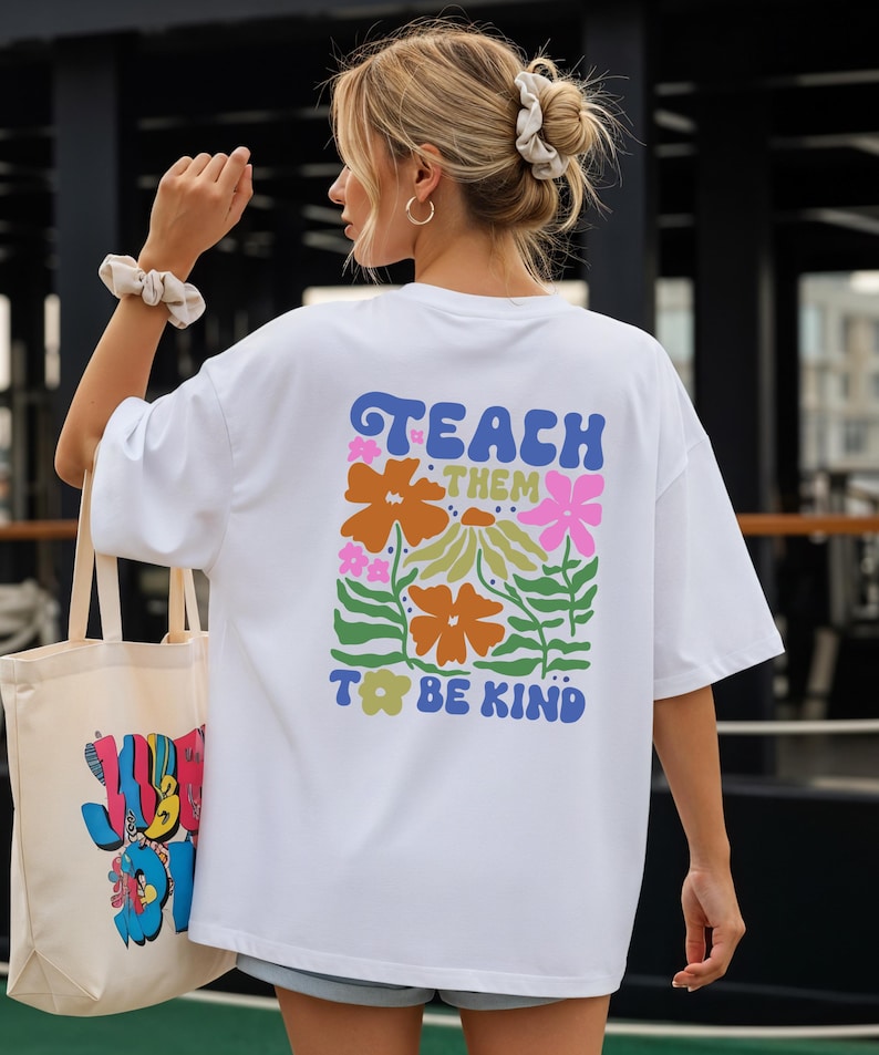 May include: White t-shirt with the text "TEACH THEM TO BE KIND" in blue, orange, and pink floral design. A tote bag with a colorful graphic is held in the left hand. The person is wearing a scrunchie and hoop earrings.