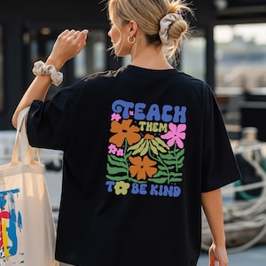 May include: Black t-shirt with a colorful floral design and the words "TEACH THEM TO BE KIND" in a retro font. The design features orange, pink, and yellow flowers with green leaves. The shirt is a casual, oversized style.