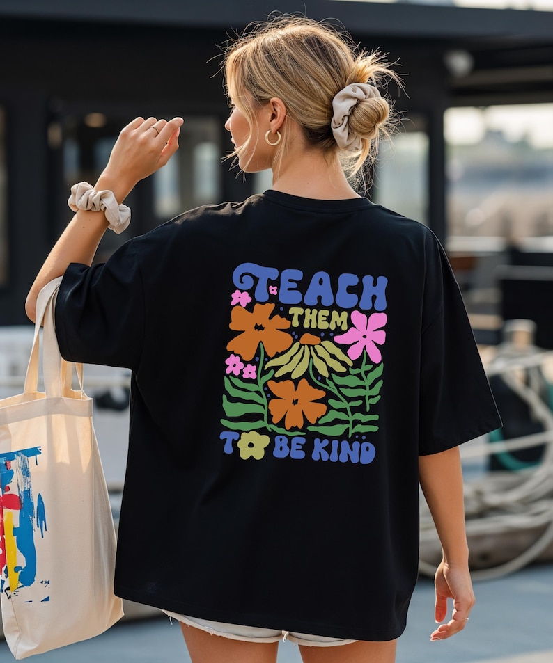 May include: Black t-shirt with a colorful floral design and the words "TEACH THEM TO BE KIND" in a retro font. The design features orange, pink, and yellow flowers with green leaves. The shirt is a casual, oversized style.