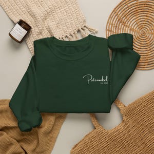 Puede incluir: Una sudadera de manga larga en color verde bosque oscuro con texto blanco en cursiva en el pecho izquierdo que dice Patenonkel Est. 2025. La prenda tiene cuello redondo, pu&ntilde;os acanalados y un acabado de color s&oacute;lido en el frente.