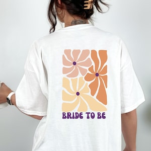 Könnte beinhalten: Weißes T-Shirt mit einem Blumendesign in Pfirsich-, Orange- und Gelbtönen. Der Text "BRIDE TO BE" ist in Lila unter den Blumen aufgedruckt. Das T-Shirt hat eine lockere Passform.