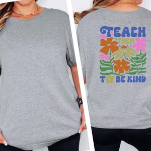 May include: A heather gray t-shirt with the words "TEACH THEM TO BE KIND" printed on the back, along with a floral design in orange, pink, and green. The shirt is a casual style.